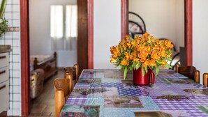 A dining table with a colorful quilted tablecloth, a red vase with orange flowers, and wooden chairs.