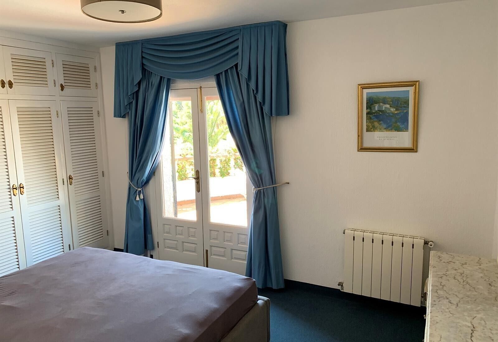 A bedroom with a bed, a radiator, a window with curtains, a door, and a framed picture on the wall.