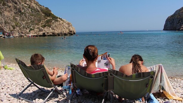 Drei Personen sitzen auf Strandstühlen an einem Kiesstrand, im Hintergrund eine felsige Klippe und ruhiges Meer.