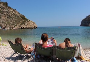 Three people seated on beach chairs on a pebbled shore, with a rocky cliff and calm sea in the background. - Villa 'Casa Pera' with Sea View, Pool, Wi-Fi, Garden & Terrace (Jávea)