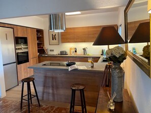 A modern kitchen with wooden cabinets, a central island, bar stools, and a large mirror. - Villa 'Casa Pera' with Sea View, Pool, Wi-Fi, Garden & Terrace (Jávea)