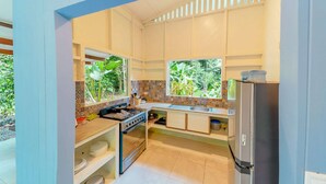Une cuisine compacte équipée d’une cuisinière à gaz, d’un évier et d’un réfrigérateur. Deux fenêtres offrent une vue sur la verdure environnante.
