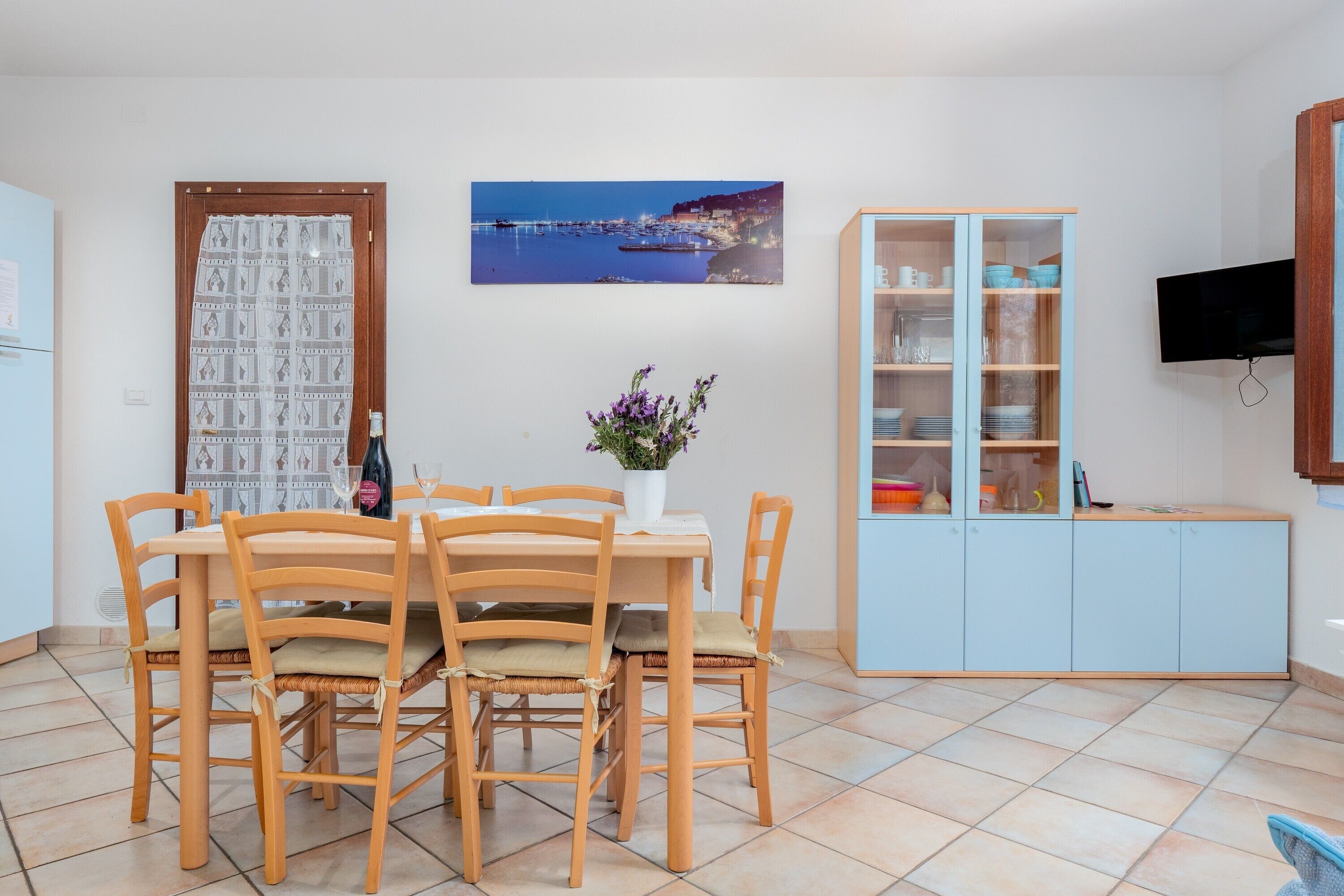 Un coin repas avec une table et des chaises en bois, un vase rempli de fleurs, un buffet contenant de la vaisselle et un téléviseur fixé au mur.