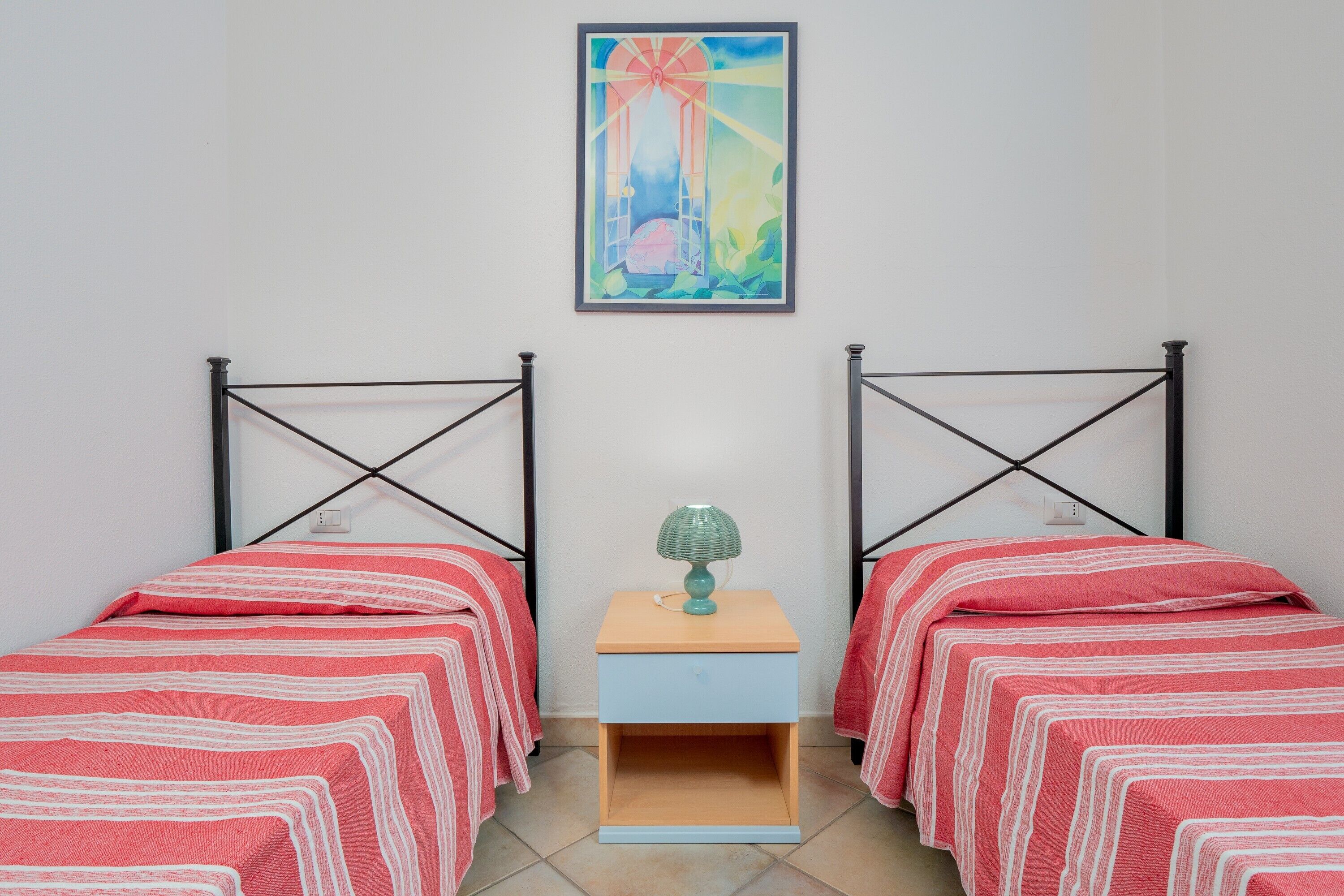 A room with two beds, a small table with a lamp, and a painting on the wall.