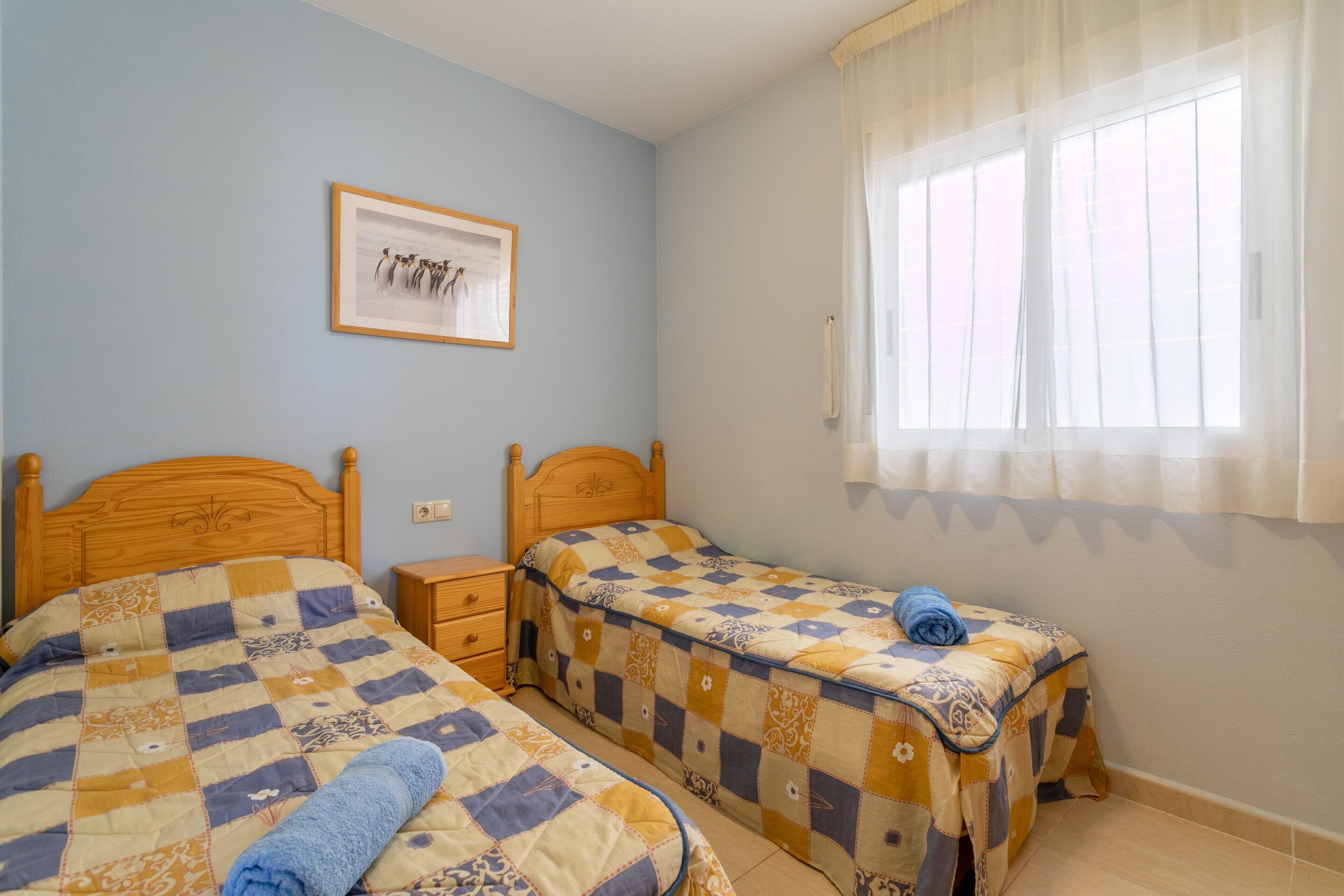 A bedroom with two beds, a nightstand, a framed picture on the wall, and a window with sheer curtains.