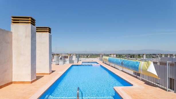 A rooftop pool with clear blue water, surrounded by a metal fence, and a view of distant mountains.