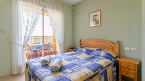 A bedroom with a bed, a wooden nightstand, a window with sheer curtains, a wooden chair, and a small table.