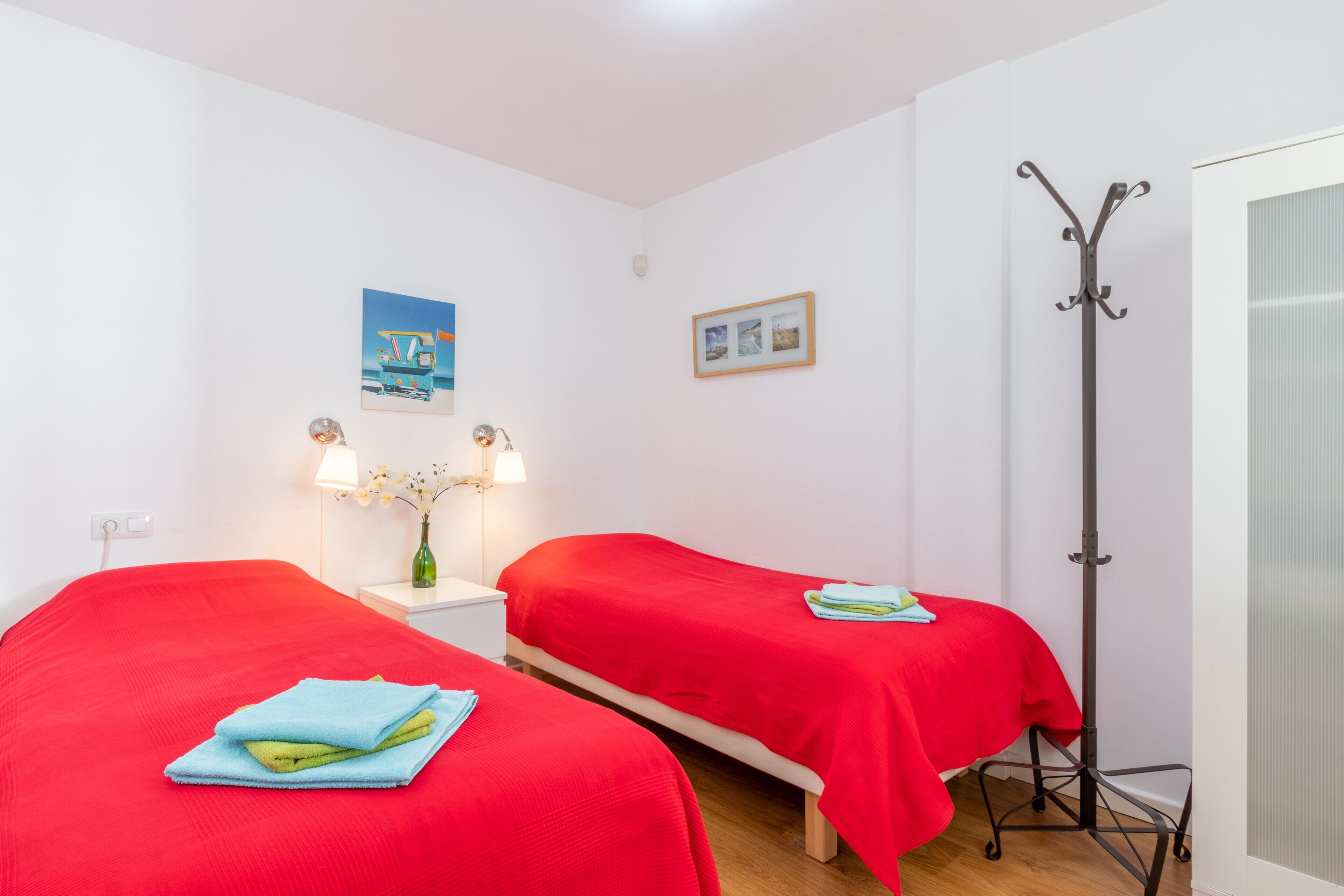 A room with two beds, a nightstand with a lamp, a picture on the wall, and a coat rack.