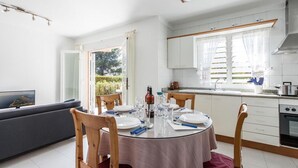 A dining area with a round table set for four, a kitchen with white cabinets, and a view of greenery outside. - Apartment 'Xiprer' with Terrace, Shared Pool & Wi-Fi (PORT POLLENÇA)