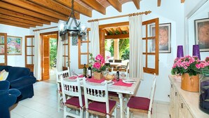 Un comedor con techo de madera, sillas blancas con cojines rojos, una mesa dispuesta para dos personas y vistas a la vegetación exterior.