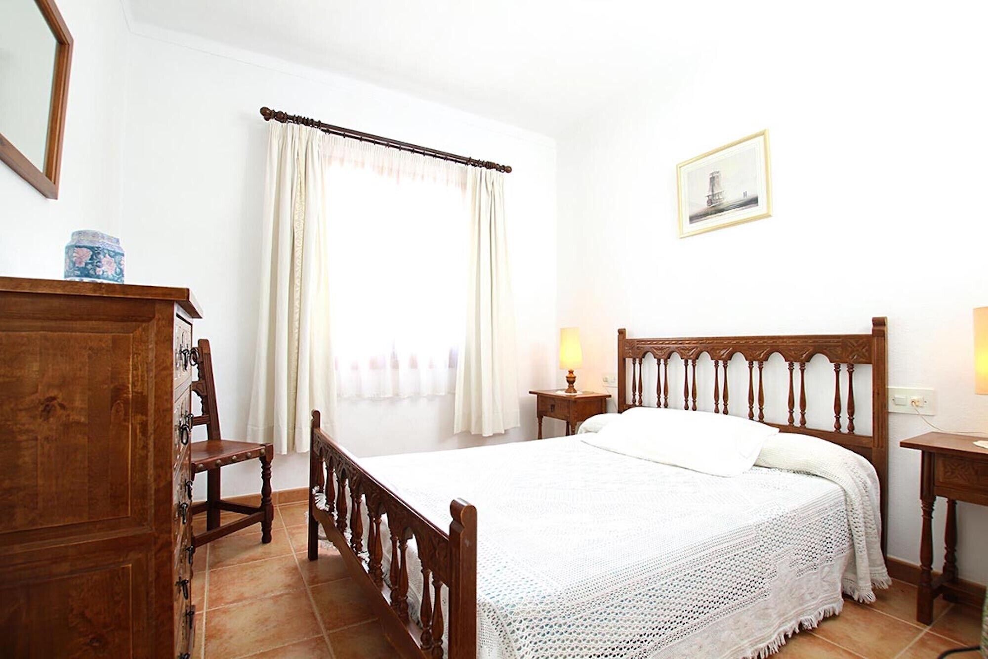 Une chambre à coucher avec un lit en bois, une table de chevet avec une lampe, une commode et une fenêtre avec des rideaux.