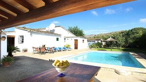 A poolside area with a wooden pergola, a table with a bowl of fruit, and a white building with a tiled roof in the background. - Country House "Casa Rei Vila" with Mountain View (Pollensa)