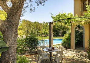 An outdoor patio with a table and chairs, a pool, and lush greenery.