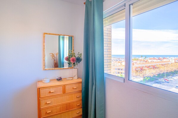 Une pièce comprenant une commode en bois, un miroir, un vase avec des fleurs et une fenêtre donnant sur des immeubles et un parking.