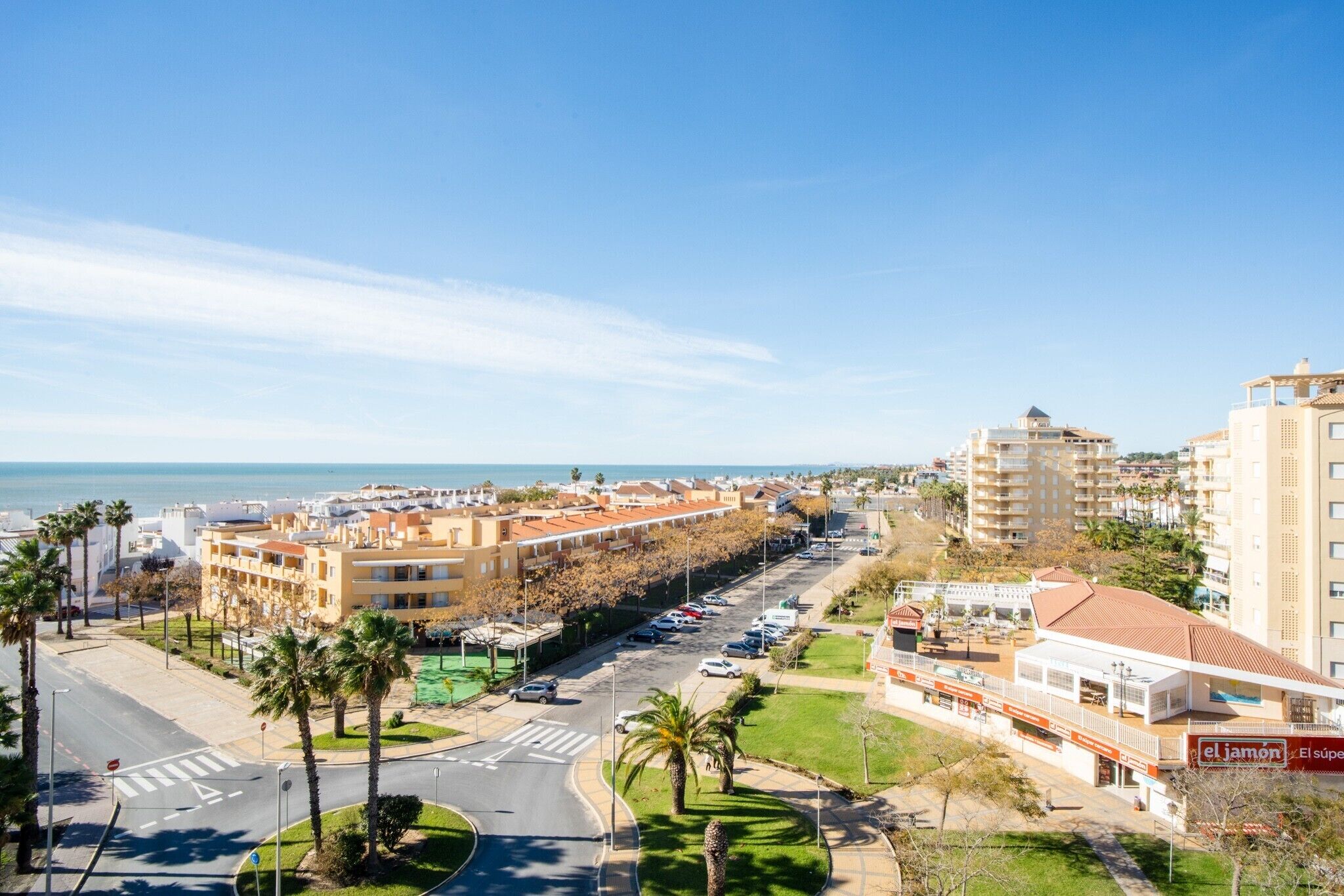 A coastal cityscape with a prominent hotel featuring a red and white facade, a parking lot, and a view of the sea.
