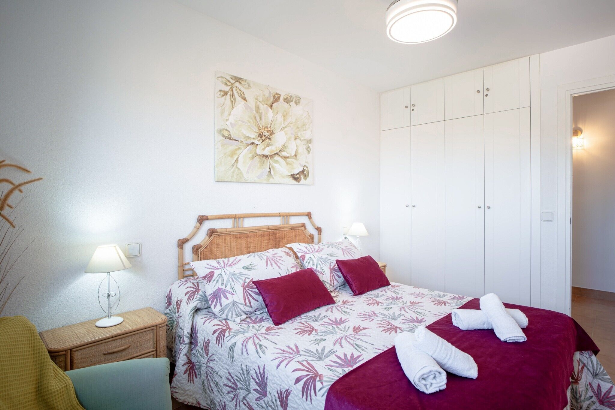 A bedroom with a bed, a nightstand, a lamp, a wardrobe, and a floral painting on the wall.