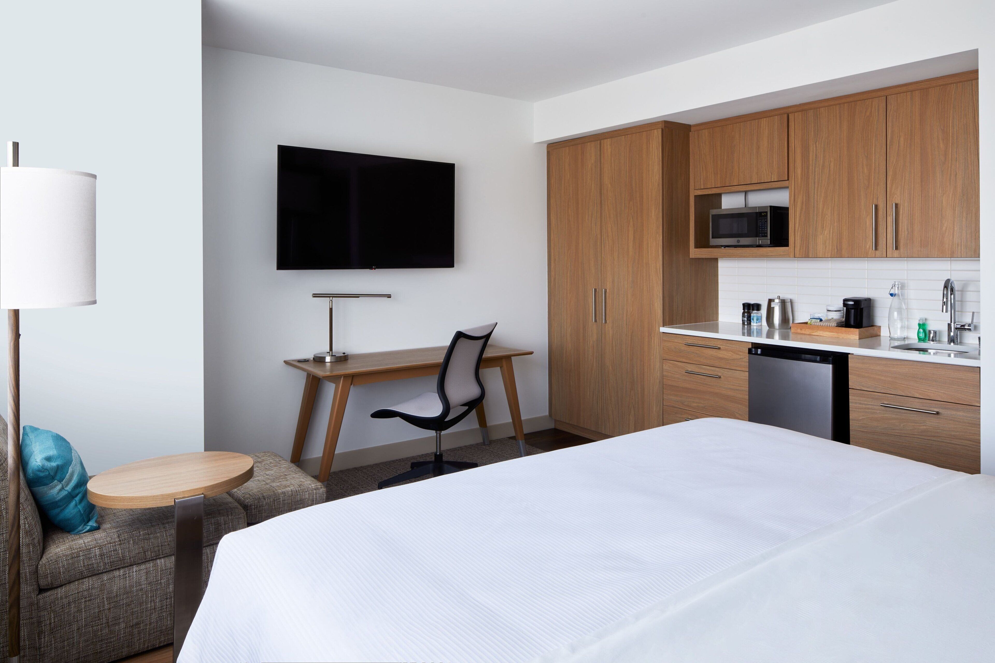 A hotel room with a bed, a desk with a chair, a television, wooden cabinets, a microwave, and a dishwasher.