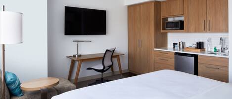 Habitación de hotel con cama, escritorio con silla, televisor, armarios de madera, microondas y lavavajillas.