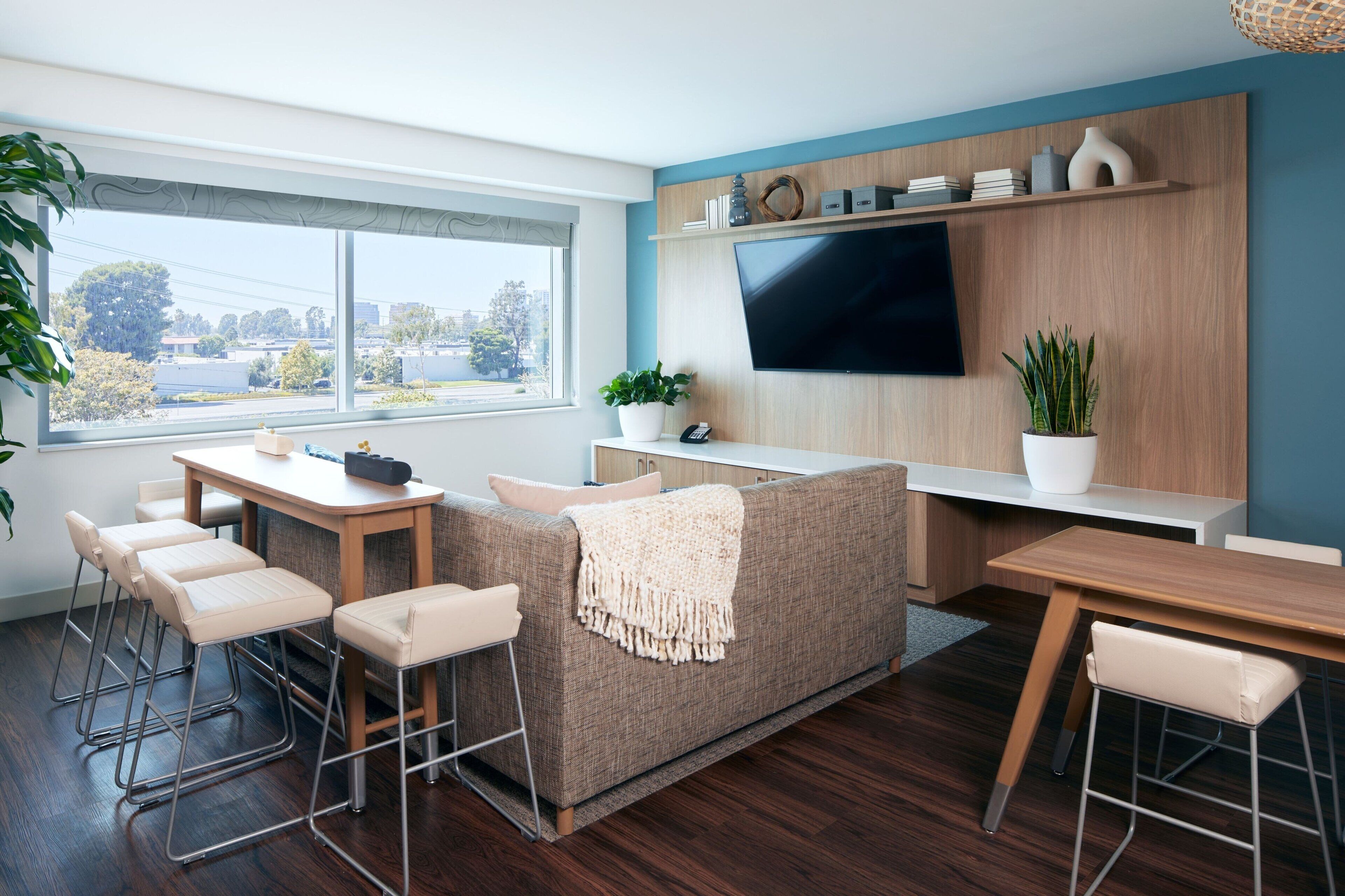 Een moderne woonkamer met een bank, salontafel en barstoelen. Er is een tv aan de muur bevestigd en een raam met uitzicht op de stad.