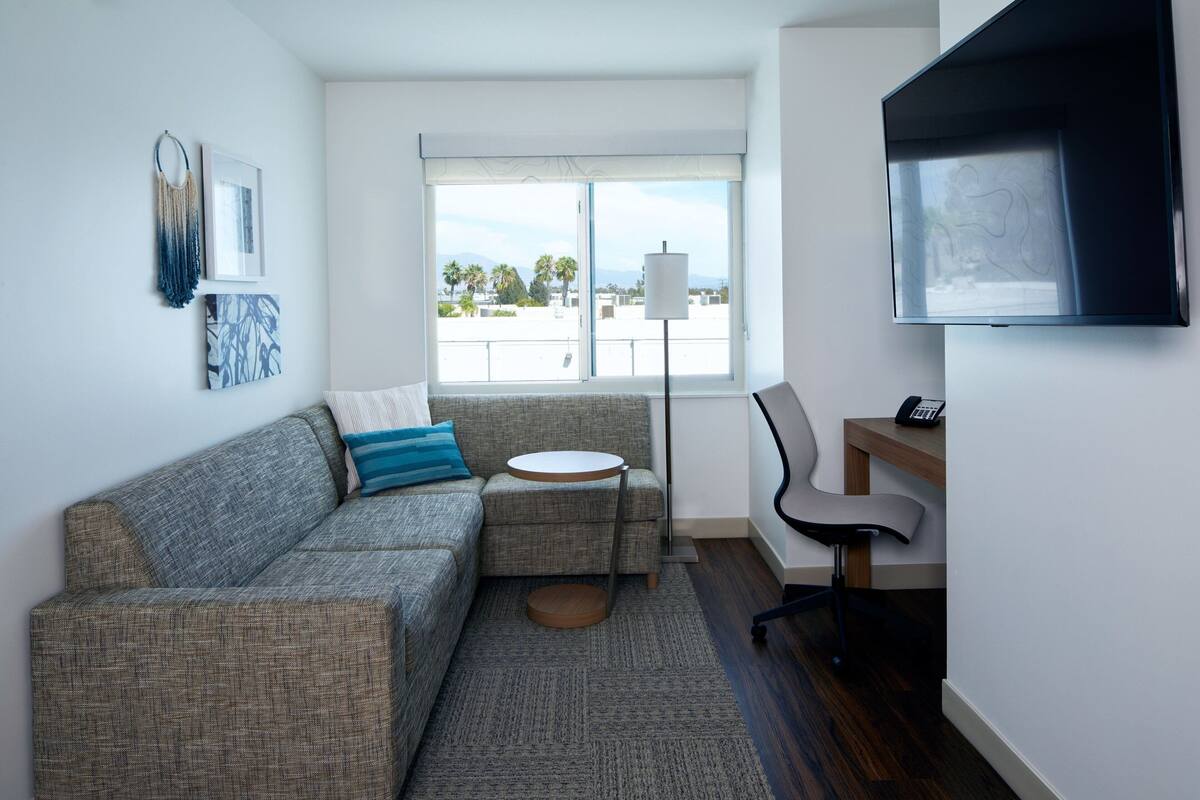 Ein modernes Wohnzimmer mit einem grauen Sofa, einem kleinen runden Tisch, einem an der Wand befestigten Fernseher und Blick ins Freie durch ein großes Fenster.