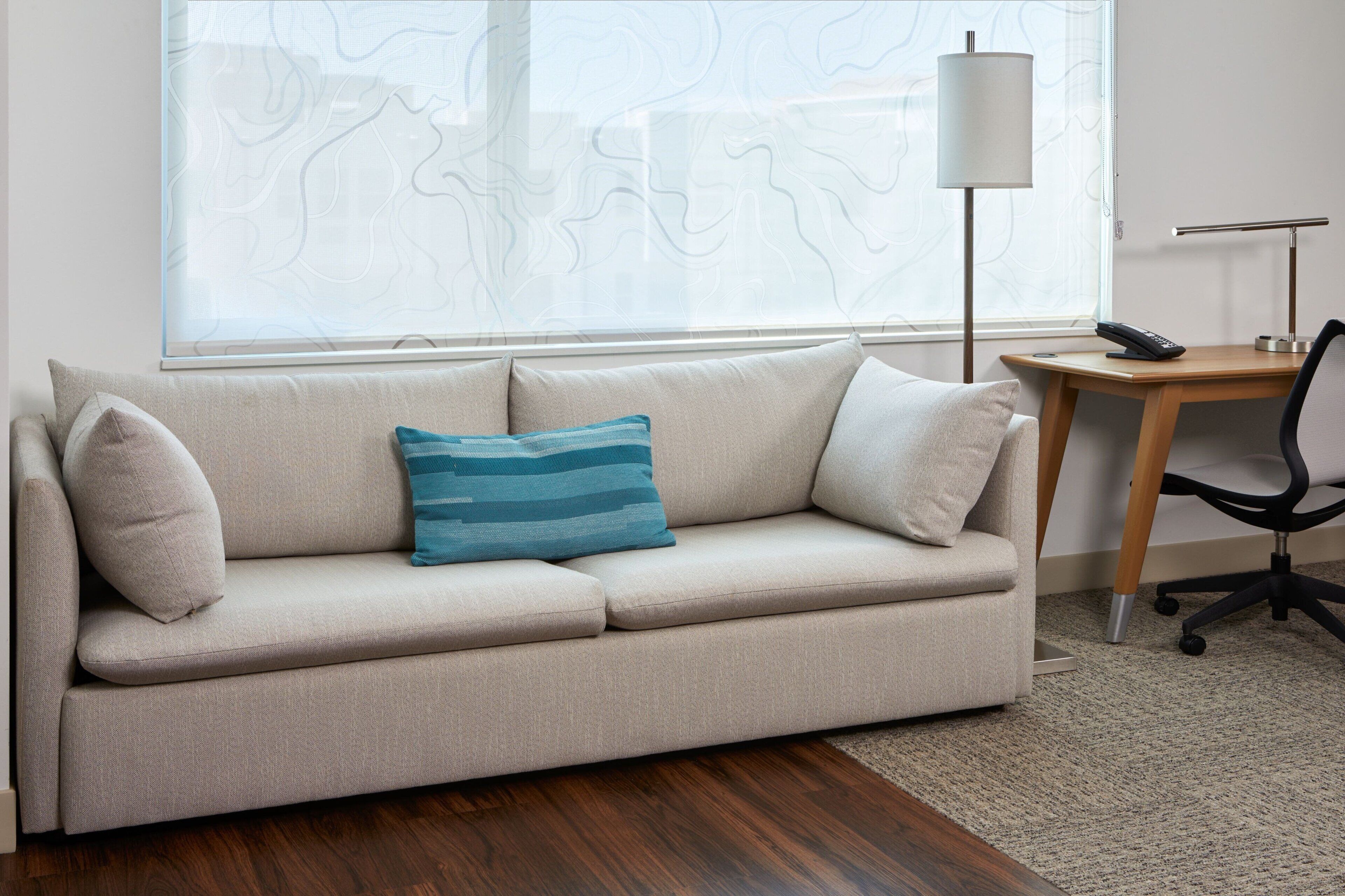 A beige sofa with a blue pillow, a wooden desk with a lamp, and a black office chair.