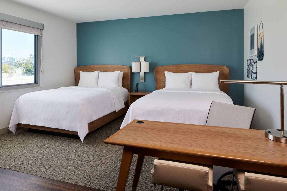Ein Hotelzimmer mit zwei Betten, einem Holztisch, einem Stuhl, einem Nachttisch und einer Lampe.