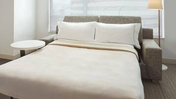 Un lit avec une couvre-lit beige, deux oreillers et une tête de lit. Une petite table ronde et une lampe se trouvent sur le côté droit.