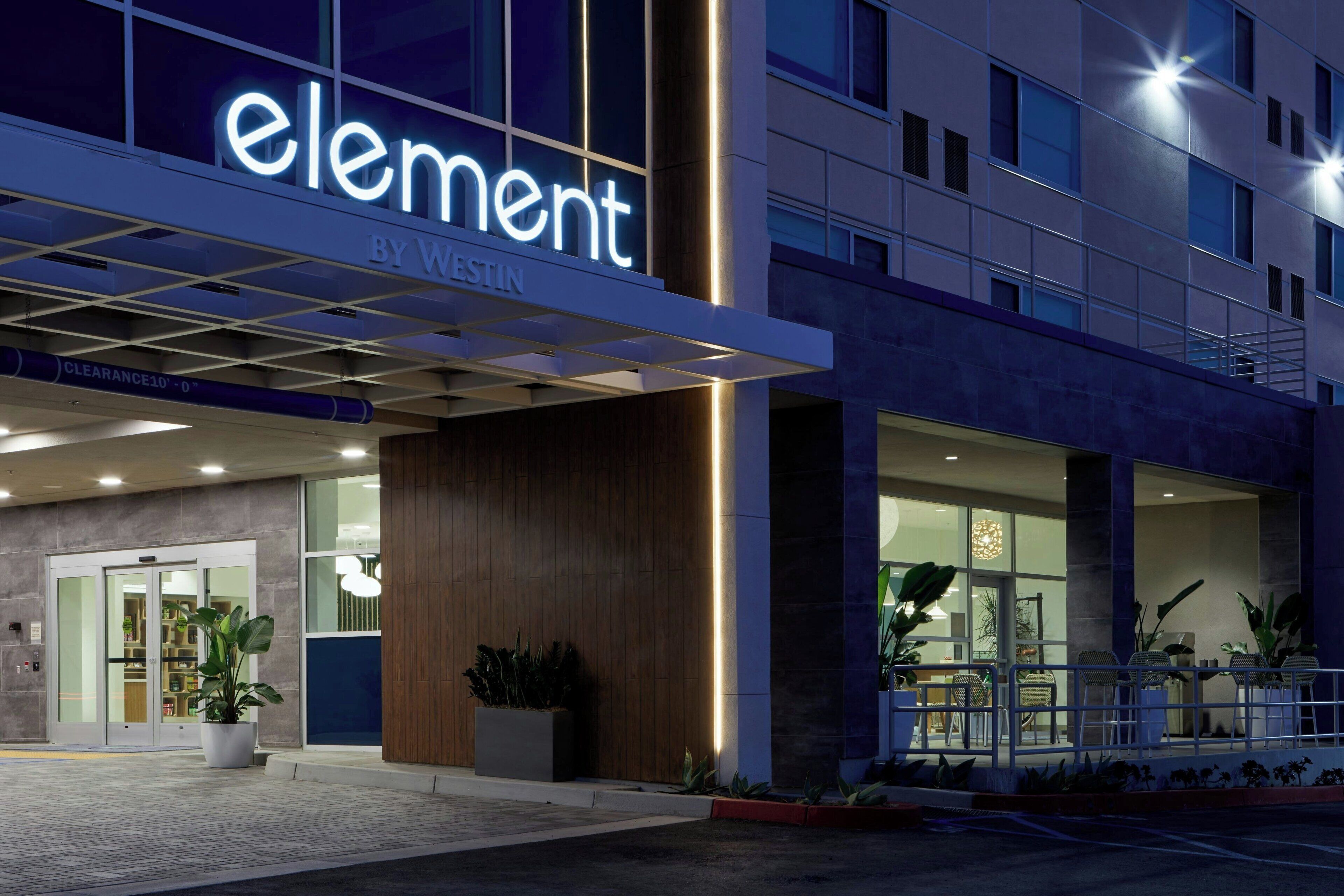 Entrada de hotel con diseño moderno, destacando un letrero grande de 'element by westin', puerta de cristal y plantas en macetas.