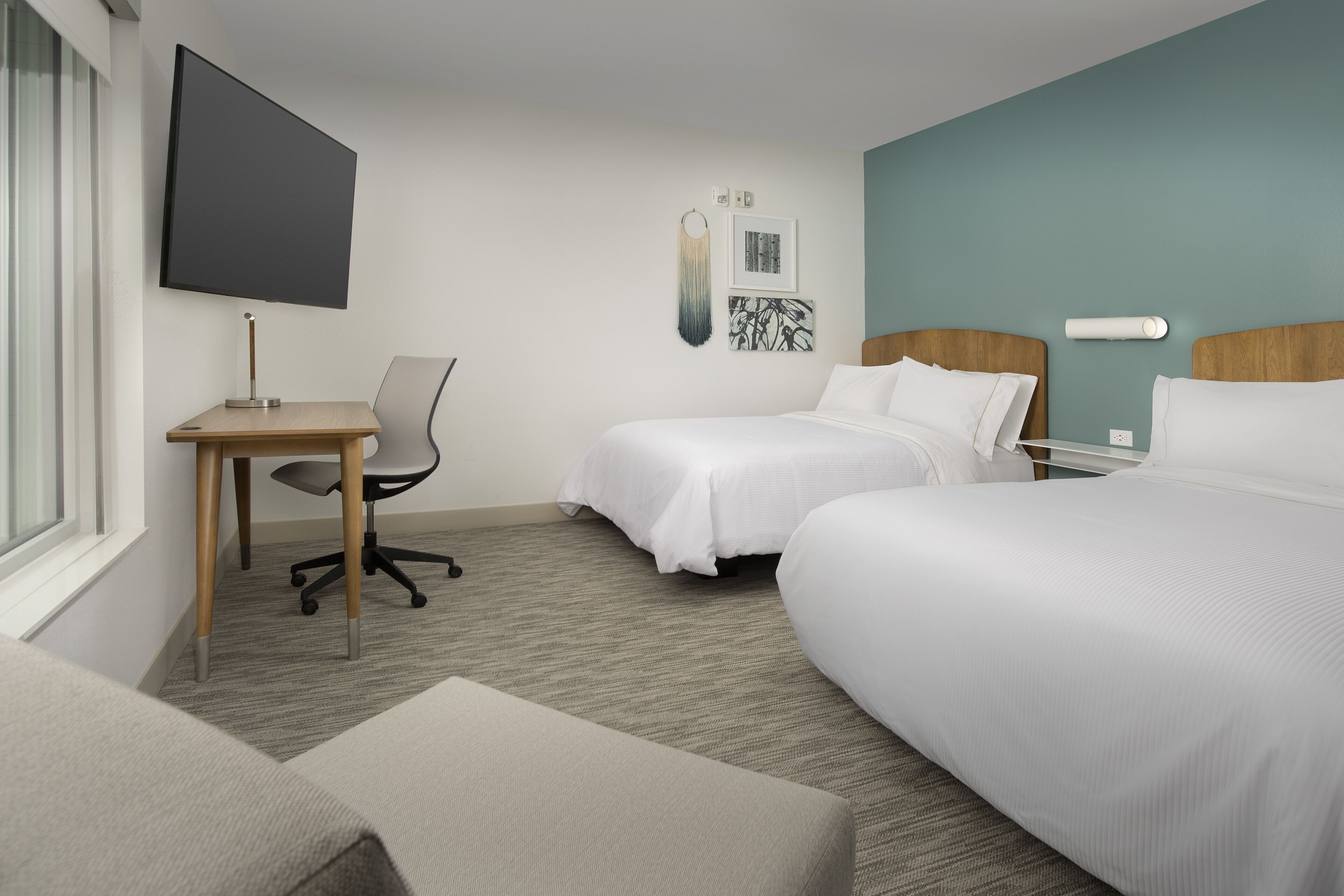 A hotel room with two beds, a desk, a chair, a TV, and artwork on the wall.