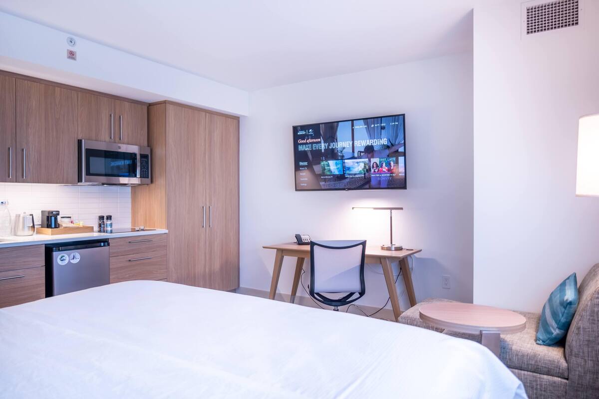 Ein modernes Hotelzimmer mit Bett, Schreibtisch, Sessel, Fernseher und einer Küchenzeile.