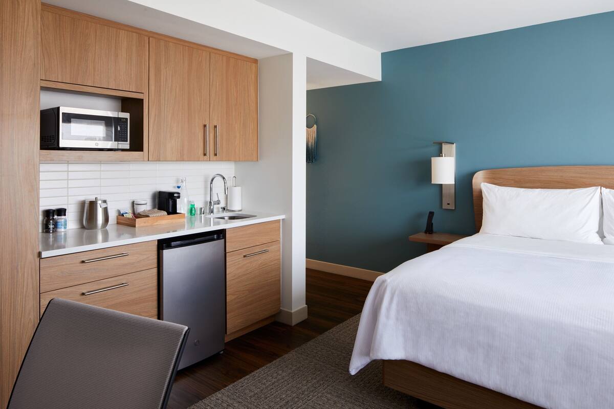 Ein modernes Hotelzimmer mit einer Holzküchenzeile, einem Bett mit weißen Leinentextilien, einer blauen Akzentwand und einem Nachttisch mit einer Lampe.