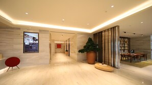 Eine moderne Hotelhalle mit Marmorboden, einem roten Sessel, einer großen Küstertopf-Pflanze, einer hölzernen Bank und einem Essbereich mit Tischen und Stühlen.