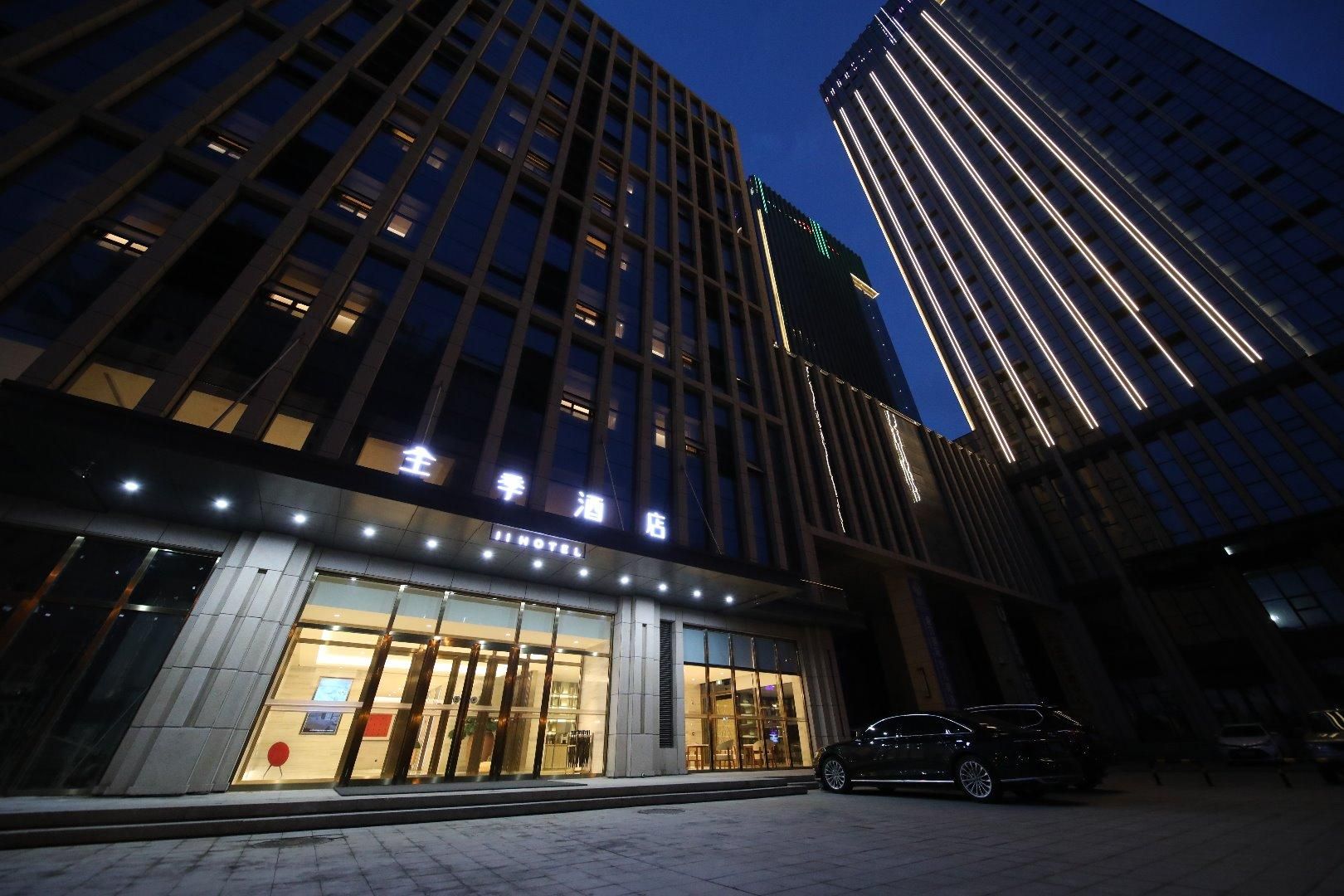 A modern hotel with a glass facade, illuminated entrance, and a parking area with cars.