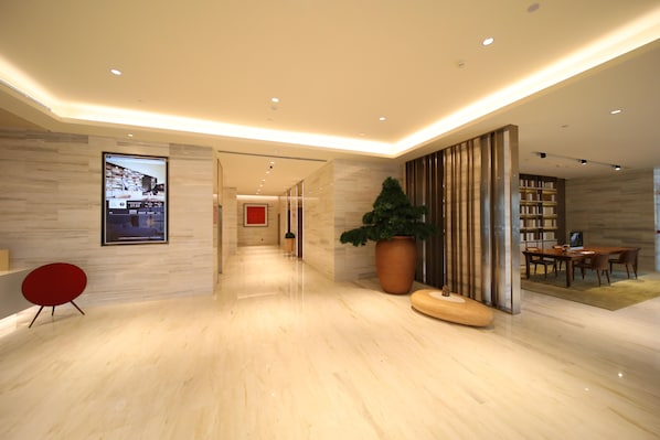 A modern hotel lobby with a marble floor, a red chair, a large potted plant, a wooden bench, and a dining area with tables and chairs. - Ji Hotel (Changchun)