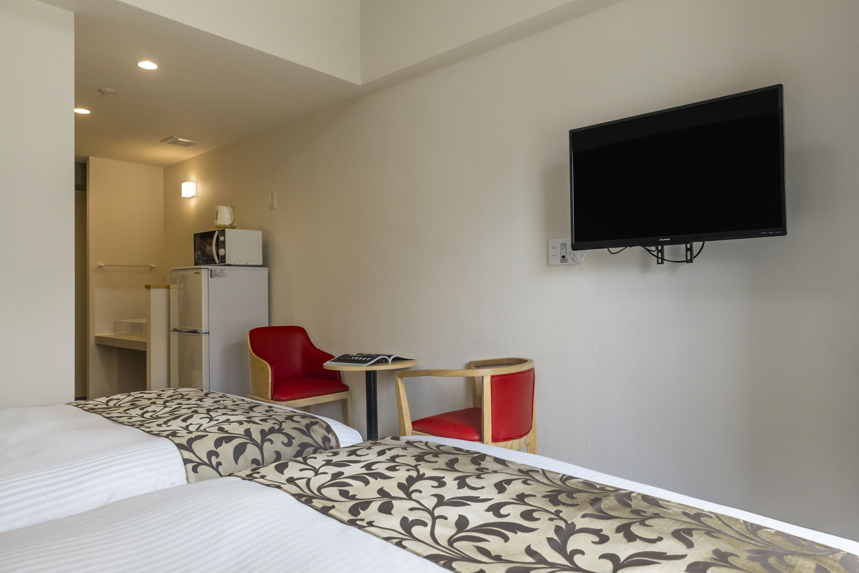 A hotel room with two beds, a TV mounted on the wall, a small table, and two red chairs.