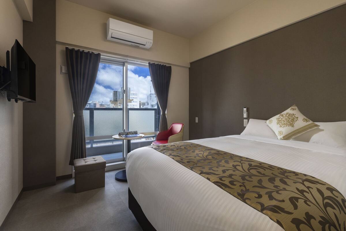 A hotel room with a large bed, a red chair, a small table, and a view of the city through a window.