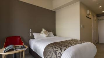 A hotel room with a bed, a red chair, a small table with a magazine, and a door.