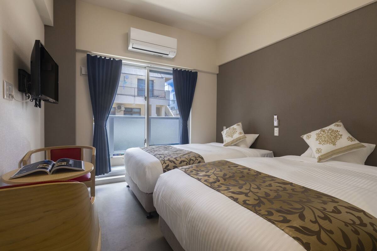 A hotel room with two beds, a TV, a desk with a book, and a window with curtains.