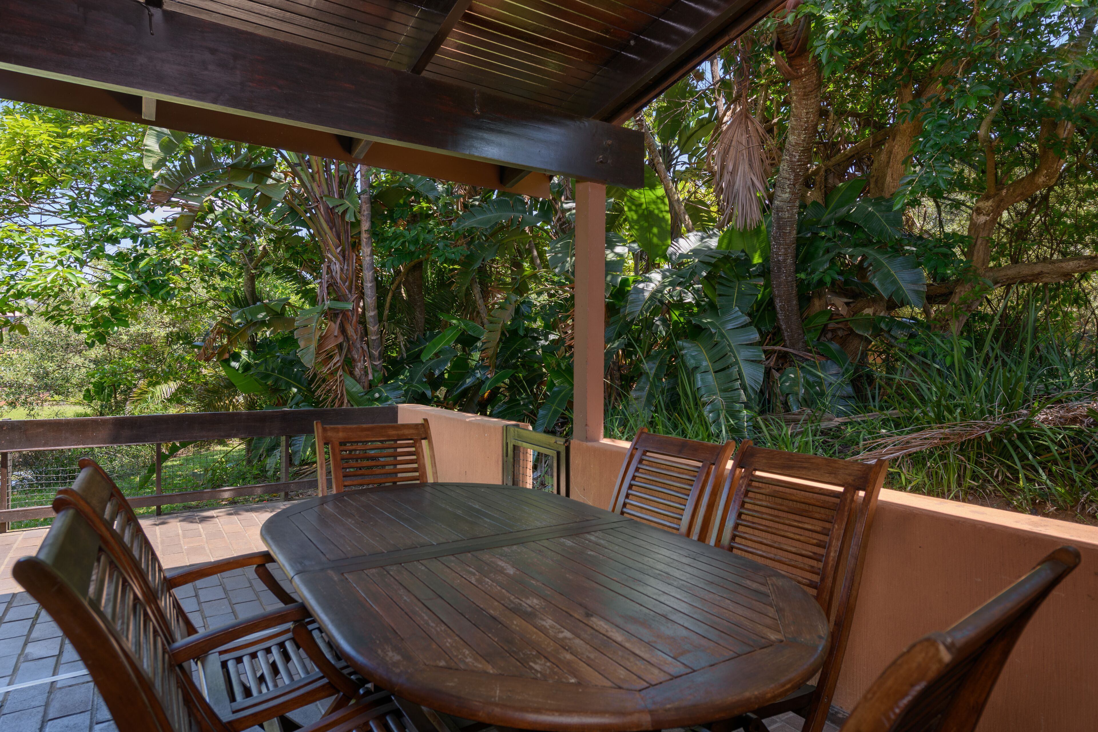Um pátio externo com mesa e cadeiras de madeira, cercado por vegetação exuberante e árvores.