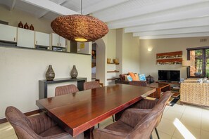 A dining area with a long wooden table, cushioned chairs, a woven pendant light, and a television in the background.