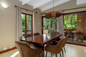 A dining area with a long wooden table, six chairs, a hanging light fixture, and large windows with curtains.