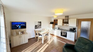 Eine moderne Küche mit weißen Schränken, eine Essecke mit einem Holztisch und Stühlen, ein schwarzes Ledersofa und ein flachbildfernseher an der Wand.
