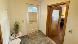 Ein Flur mit einer weißen Tür, einem Fenster mit Jalousien, einem kleinen Tisch mit Pflanze und einem Fliesenboden.
