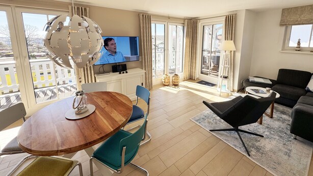 Ein modernes Wohnzimmer mit einem Holzesstisch, blauen Stühlen, einem schwarzen Sofa und einem Fernseher auf einem weißen Schrank.