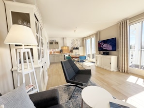 En moderne stue med sofa, sofabord og fjernsyn. Der er et køkkenområde med skabe og et spisebord.