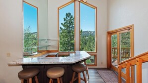 Una cocina con una isla grande, taburetes de bar y vistas a un bosque a través de ventanales altos.
