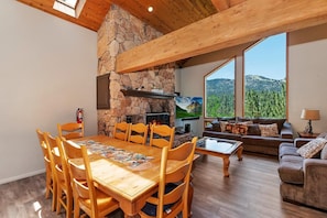 A spacious living area with a stone fireplace, wooden dining table, and a sofa facing a large window with a mountain view. - Breathtaking Ski Slope Views in Upper Moonridge! HOT TUB! Pet friendly! Arcade Games! Foosball! (Big Bear Lake)