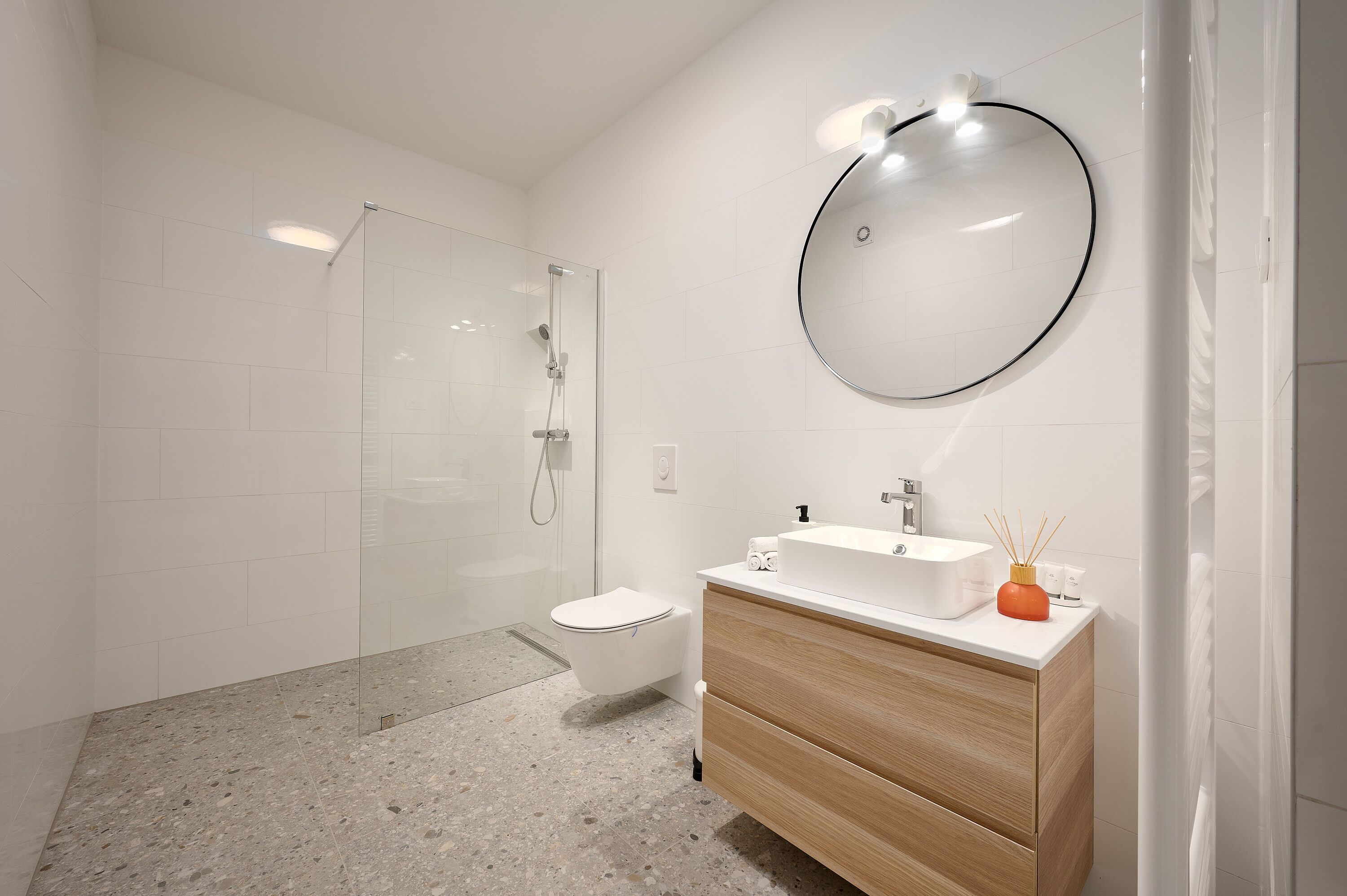 A modern bathroom with a glass-enclosed shower, a white toilet, a wooden vanity with a sink, and a round mirror.