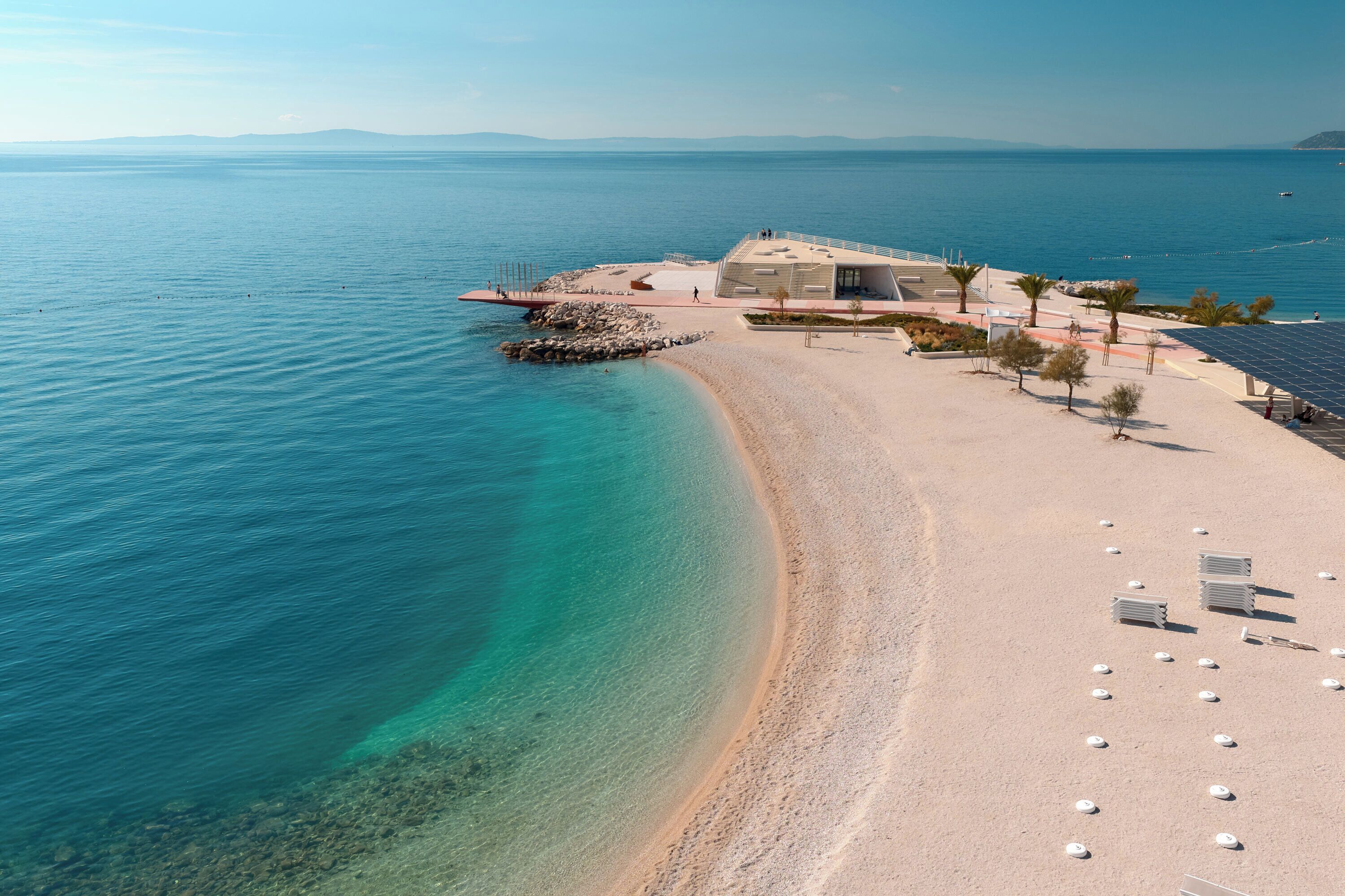 Vista aérea de una playa con aguas turquesas cristalinas, una orilla arenosa y un edificio moderno de techos planos y grandes ventanales.