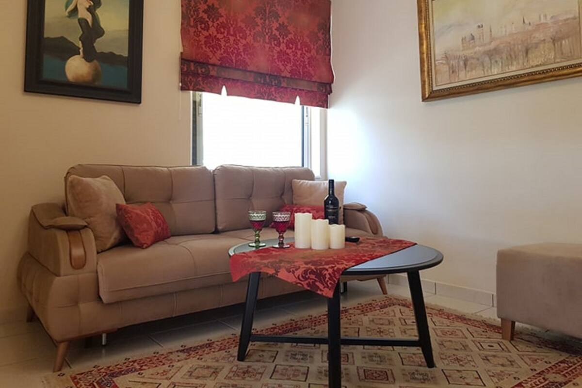 A living room with a beige sofa, a glass coffee table with a red tablecloth, a red patterned drape, and a framed painting on the wall.